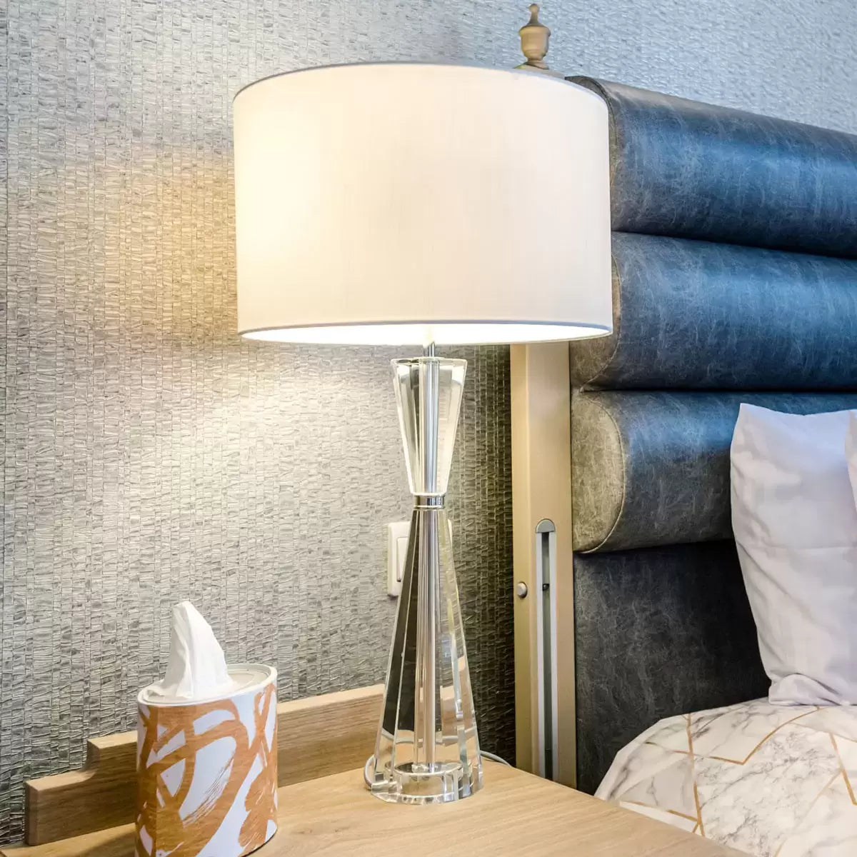 Luxurious Risha table lamp featuring a heavy, faceted hourglass-shaped crystal base, polished nickel hardware, and an elegant ivory faux silk drum shade styled in a contemporary living room interior.