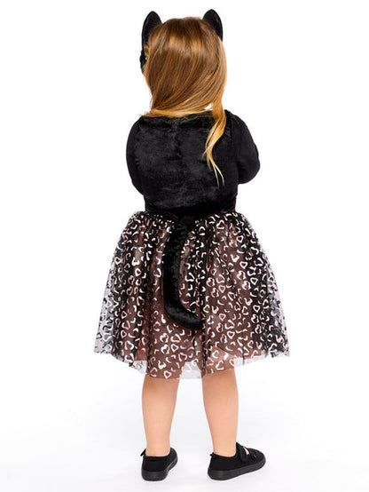 Front view of a toddler wearing the Cutie Cat costume with black velvet top, smiling cat face emblem, net leopard print skirt, attached tail, and cat ear headband in a cozy living room setting.