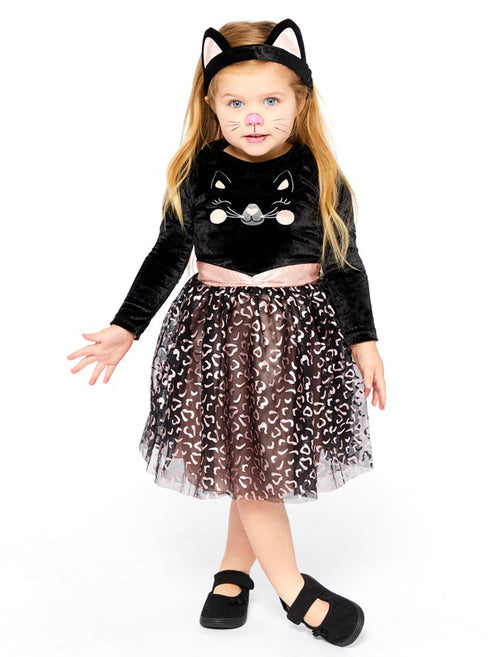 Front view of a toddler wearing the Cutie Cat costume with black velvet top, smiling cat face emblem, net leopard print skirt, attached tail, and cat ear headband in a cozy living room setting.