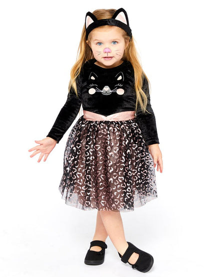 Front view of a toddler wearing the Cutie Cat costume with black velvet top, smiling cat face emblem, net leopard print skirt, attached tail, and cat ear headband in a cozy living room setting.