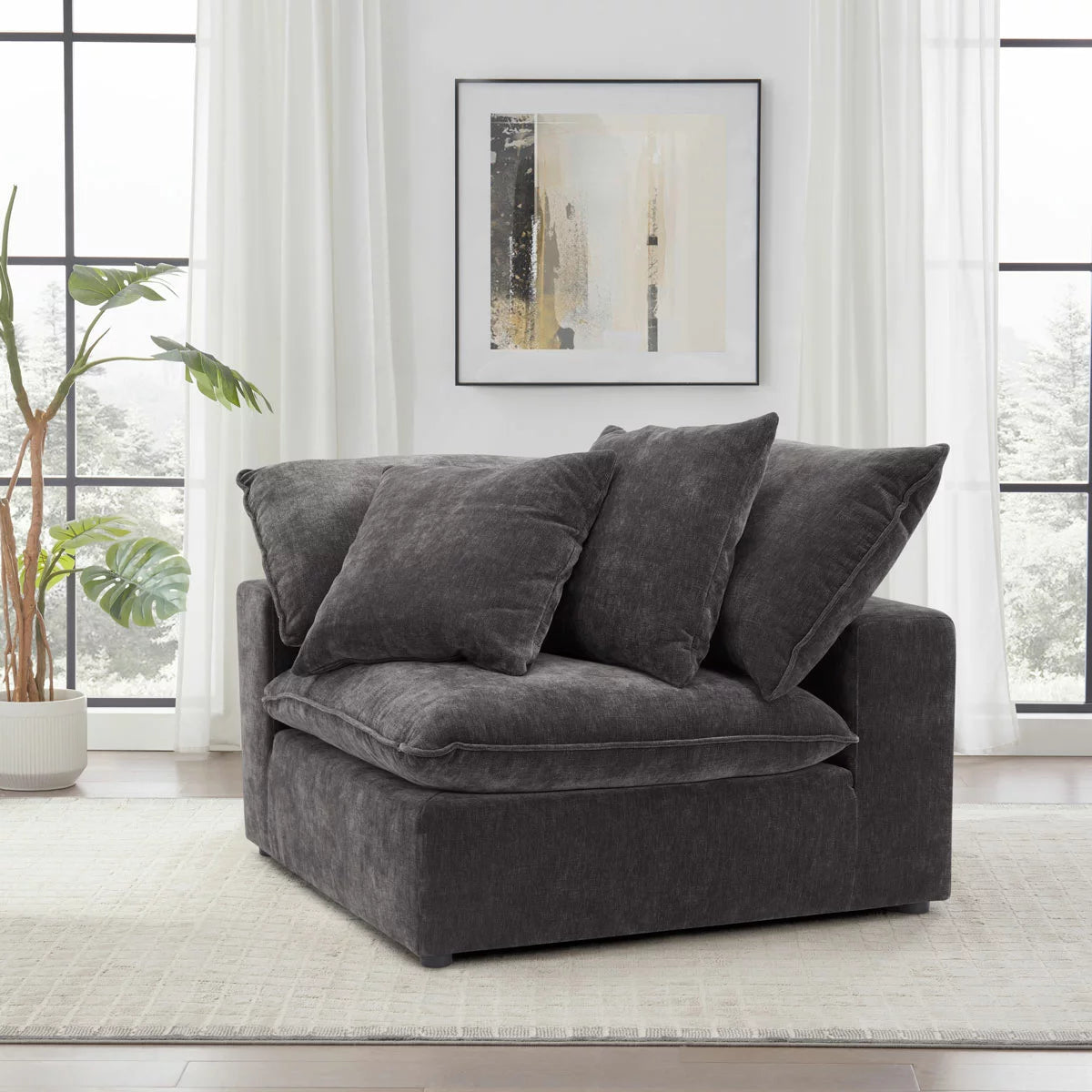 A close-up shot of the Aiden & Ivy Sereen dark grey fabric corner chair in a bright, modern living room, highlighting its textured charcoal upholstery and two matching feather-filled cushions.