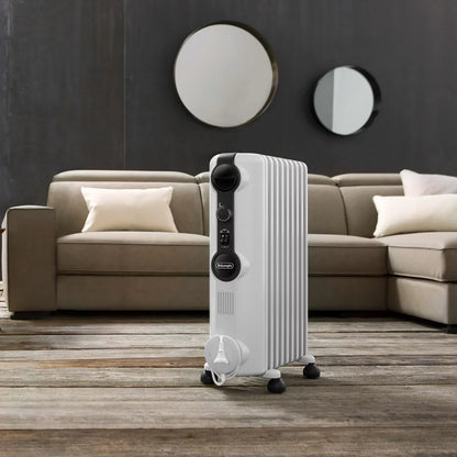 White De'Longhi Radia S 1.5kW oil filled radiator standing on a wooden floor in a modern living room setting showing pre-assembled castors and control knobs.