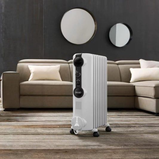 White De'Longhi Radia S 1.5kW oil filled radiator standing on a wooden floor in a modern living room setting showing pre-assembled castors and control knobs.