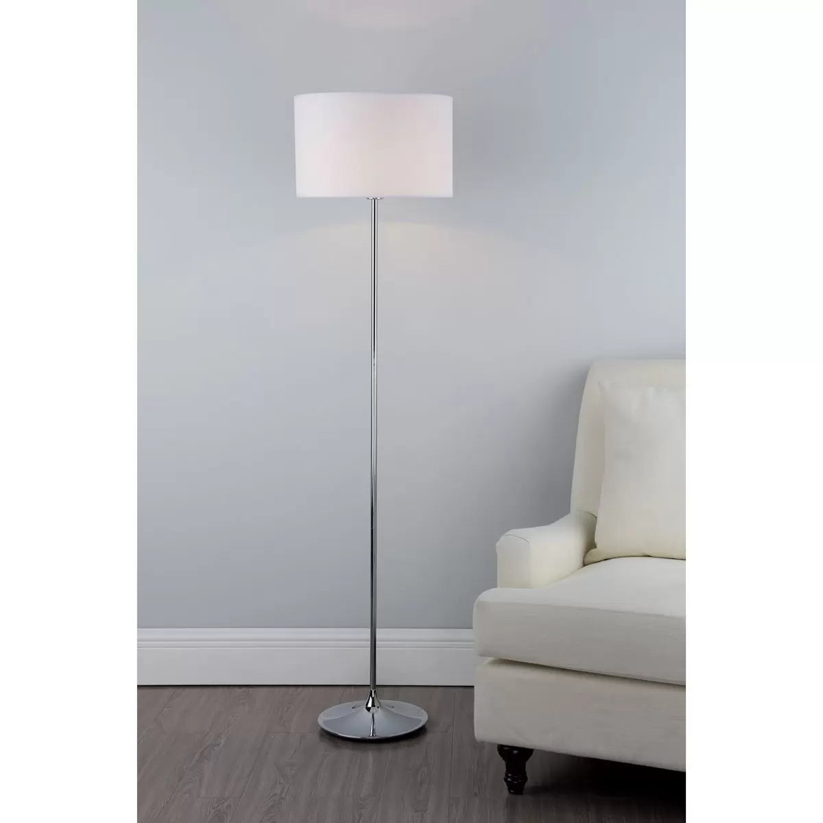 A side-angle shot of the Delta floor lamp featuring a polished chrome tear-shaped base and a clean ivory cotton drum shade casting a soft, warm glow in a contemporary living room corner.