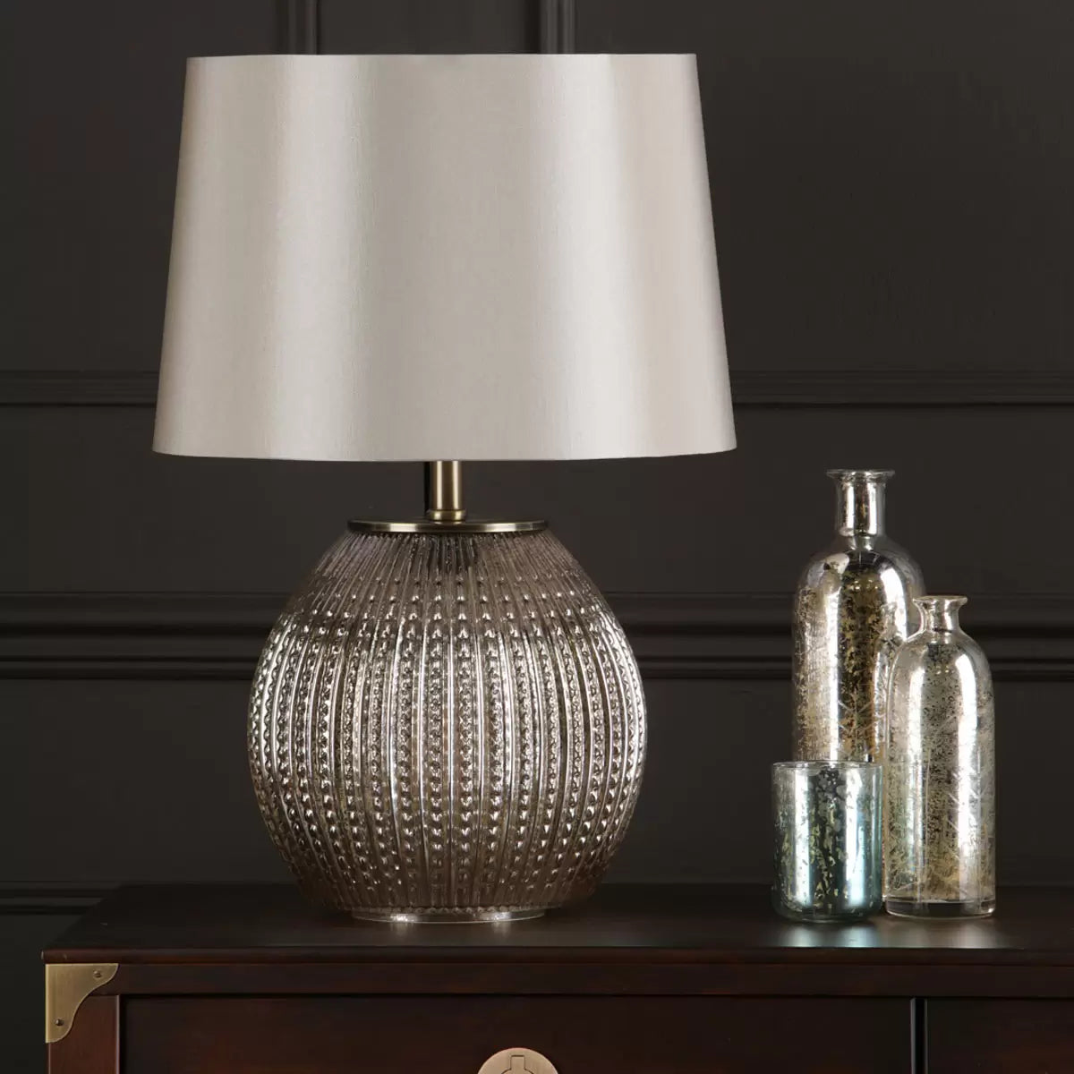 Sonia dual light table lamp featuring a glowing antique silver glass base and a warm taupe tapered fabric shade placed on a dark wood living room side table.