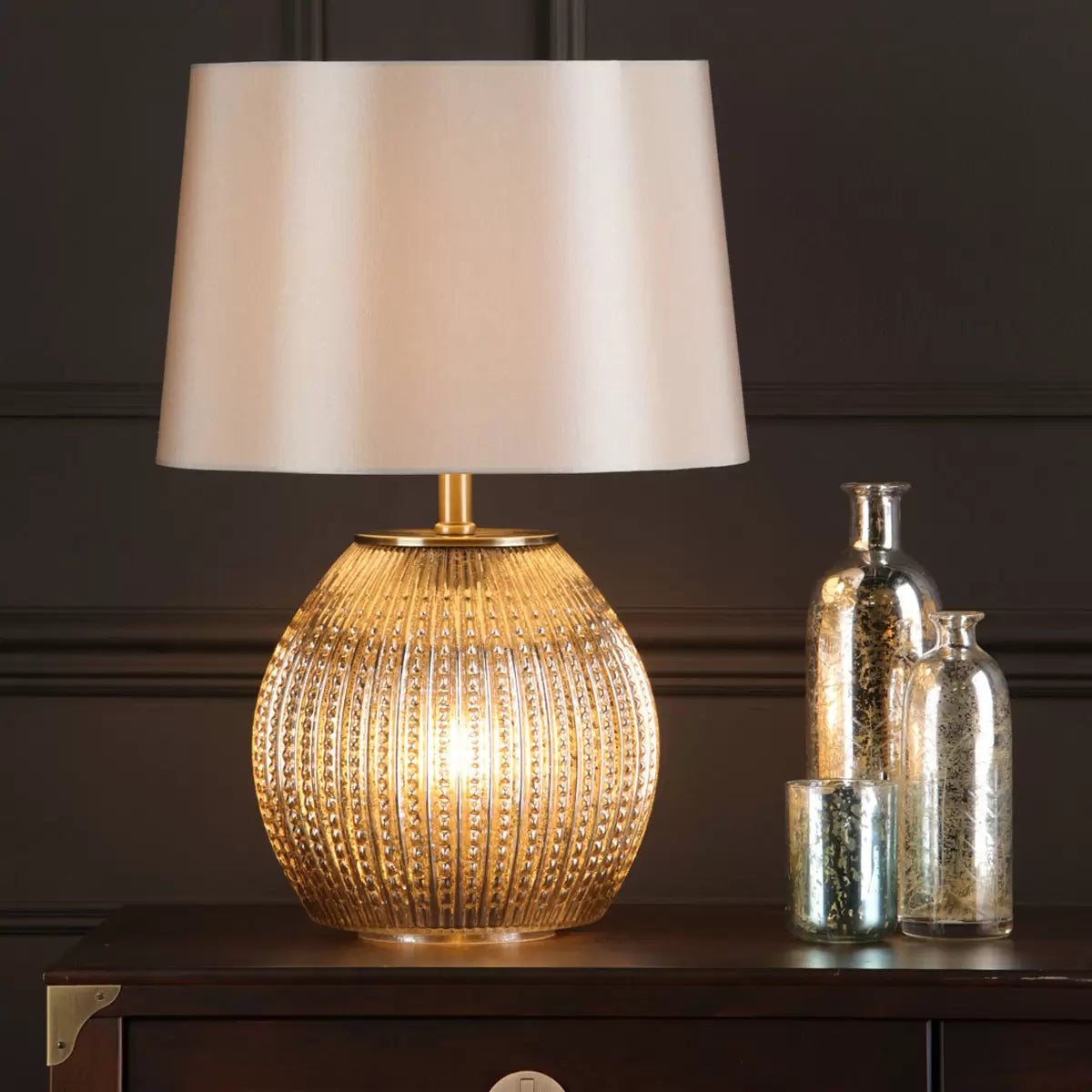 Sonia dual light table lamp featuring a glowing antique silver glass base and a warm taupe tapered fabric shade placed on a dark wood living room side table.