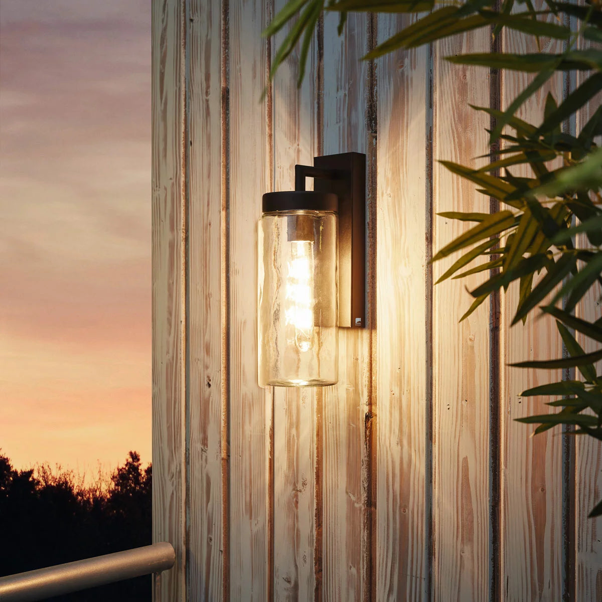 EGLO Bovolone exterior wall lamp in a matte black finish, featuring a clear cylindrical glass shade mounted on an outdoor wall at dusk.