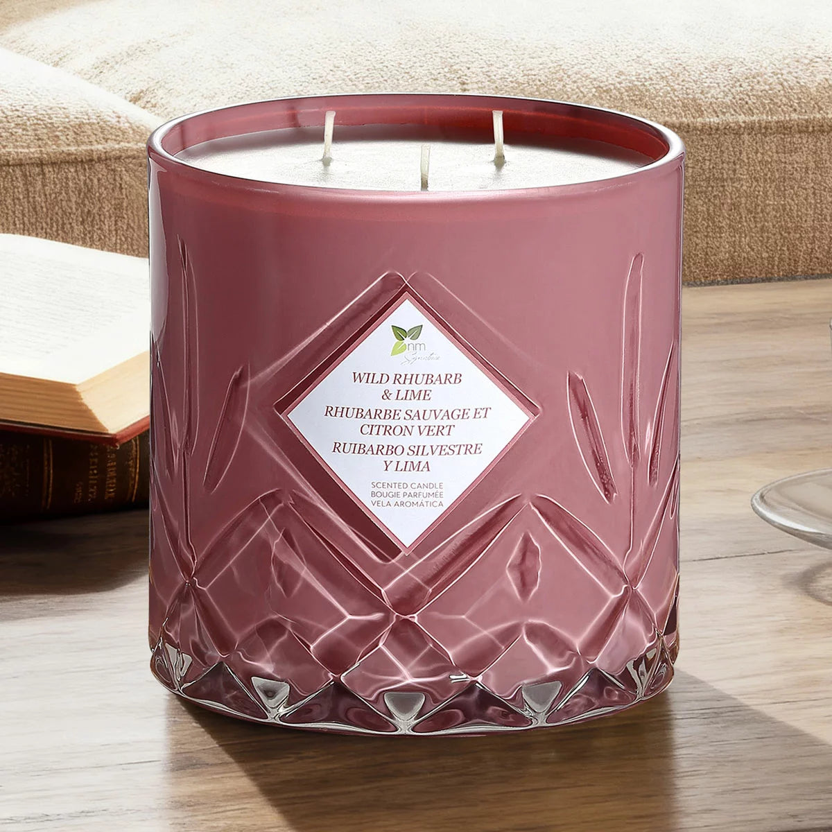 A heavy 1.58kg Nature's Mark glass candle with three lit cotton wicks glowing warmly on a rustic wooden table in a softly lit living room setting.
