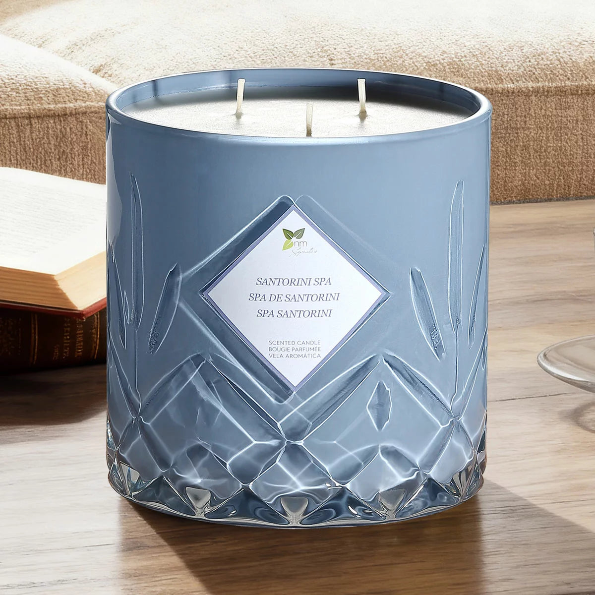 A heavy 1.58kg Nature's Mark glass candle with three lit cotton wicks glowing warmly on a rustic wooden table in a softly lit living room setting.