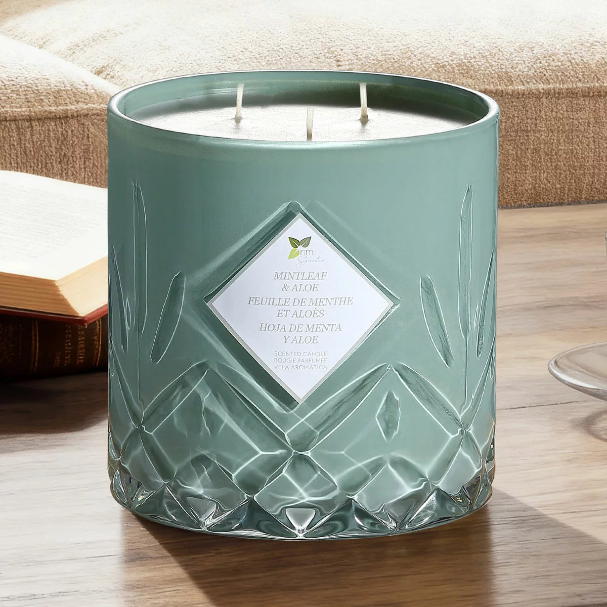 A heavy 1.58kg Nature's Mark glass candle with three lit cotton wicks glowing warmly on a rustic wooden table in a softly lit living room setting.