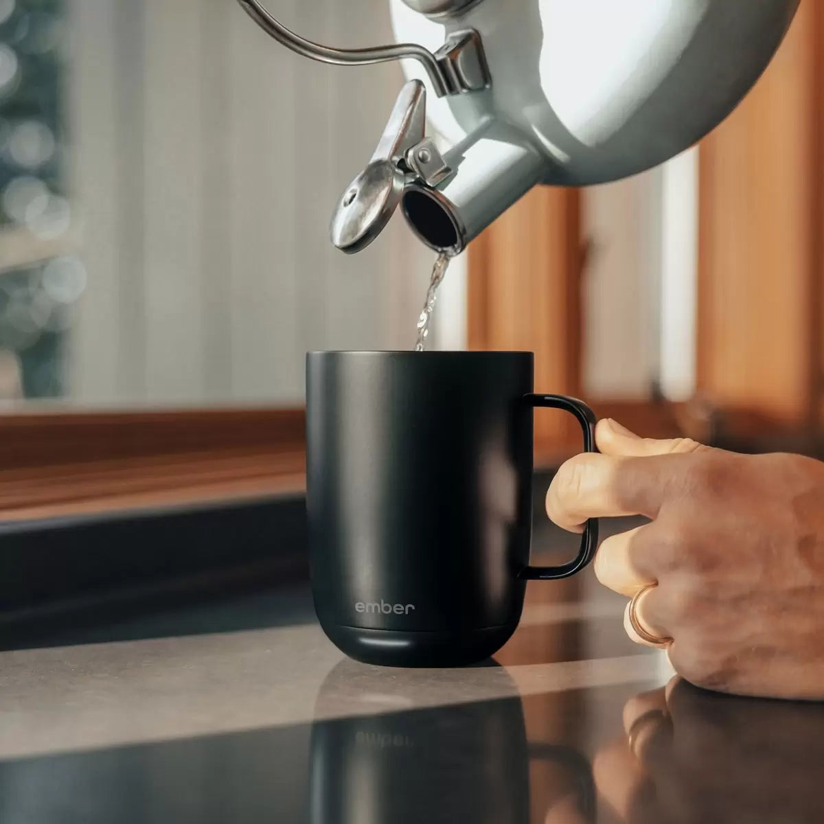 Matte black Ember Mug 2 ceramic smart mug sitting on its charging coaster next to a laptop on a modern wooden desk setting.