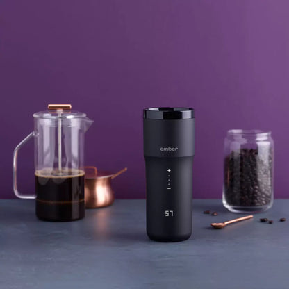 Matte black Ember Temperature Control Travel Mug 2+ with illuminated digital touch display resting on its charging coaster on a wooden office desk.