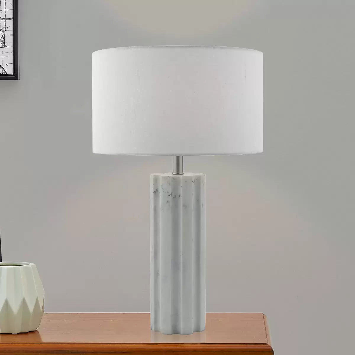 Front view of the Erebus table lamp showcasing its cylinder-shaped matte white marble effect resin base, polished chrome metal collar, and white linen drum shade on a glass desk.