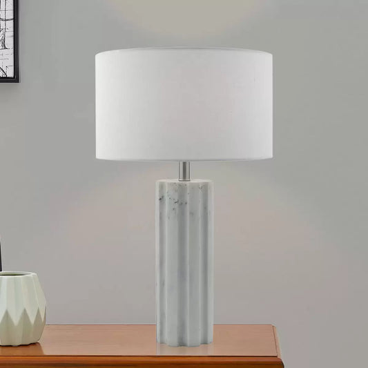 Front view of the Erebus table lamp showcasing its cylinder-shaped matte white marble effect resin base, polished chrome metal collar, and white linen drum shade on a glass desk.