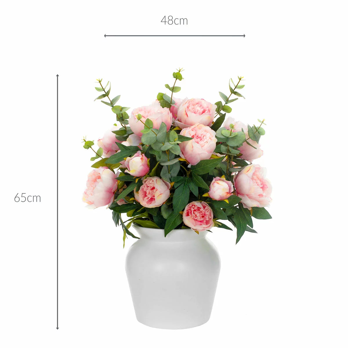 Lush artificial pink peony blooms and green eucalyptus leaves arranged inside a clear glass cylinder vase sitting on a light wooden table.