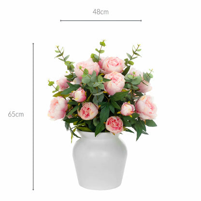 Lush artificial pink peony blooms and green eucalyptus leaves arranged inside a clear glass cylinder vase sitting on a light wooden table.