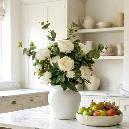 Lush artificial pink peony blooms and green eucalyptus leaves arranged inside a clear glass cylinder vase sitting on a light wooden table.