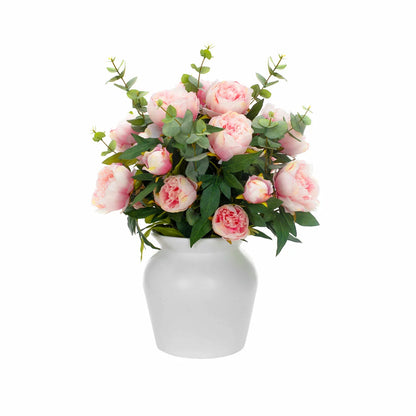 Lush artificial pink peony blooms and green eucalyptus leaves arranged inside a clear glass cylinder vase sitting on a light wooden table.