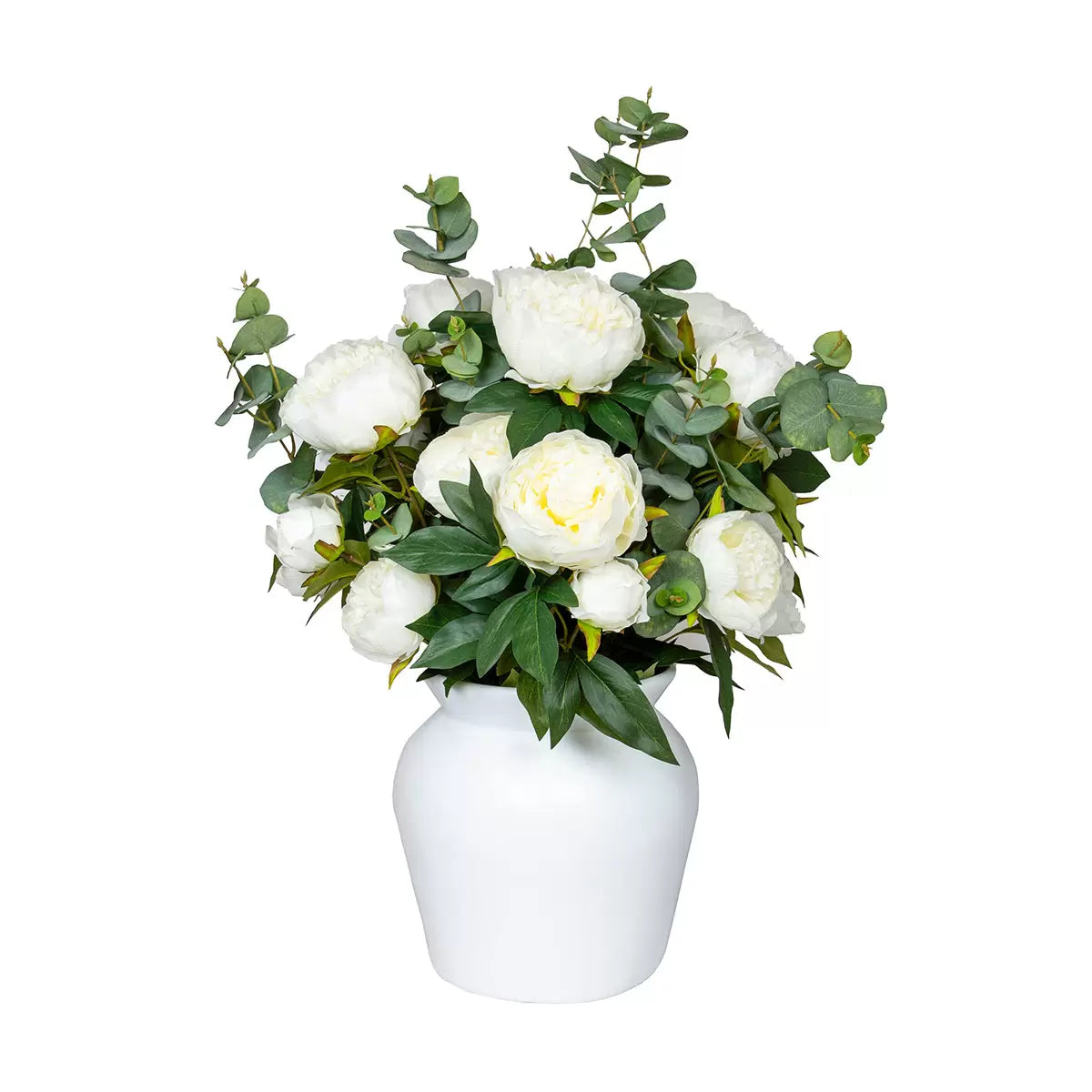 Lush artificial pink peony blooms and green eucalyptus leaves arranged inside a clear glass cylinder vase sitting on a light wooden table.