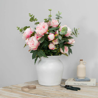 Lush artificial pink peony blooms and green eucalyptus leaves arranged inside a clear glass cylinder vase sitting on a light wooden table