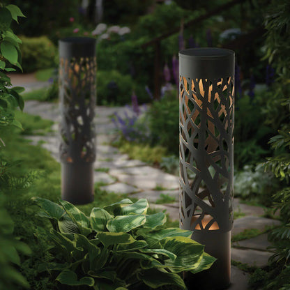 A pair of GTX steel solar bollard lights installed along a garden walkway, featuring intricate cut-out patterns that cast decorative shadows on the ground.