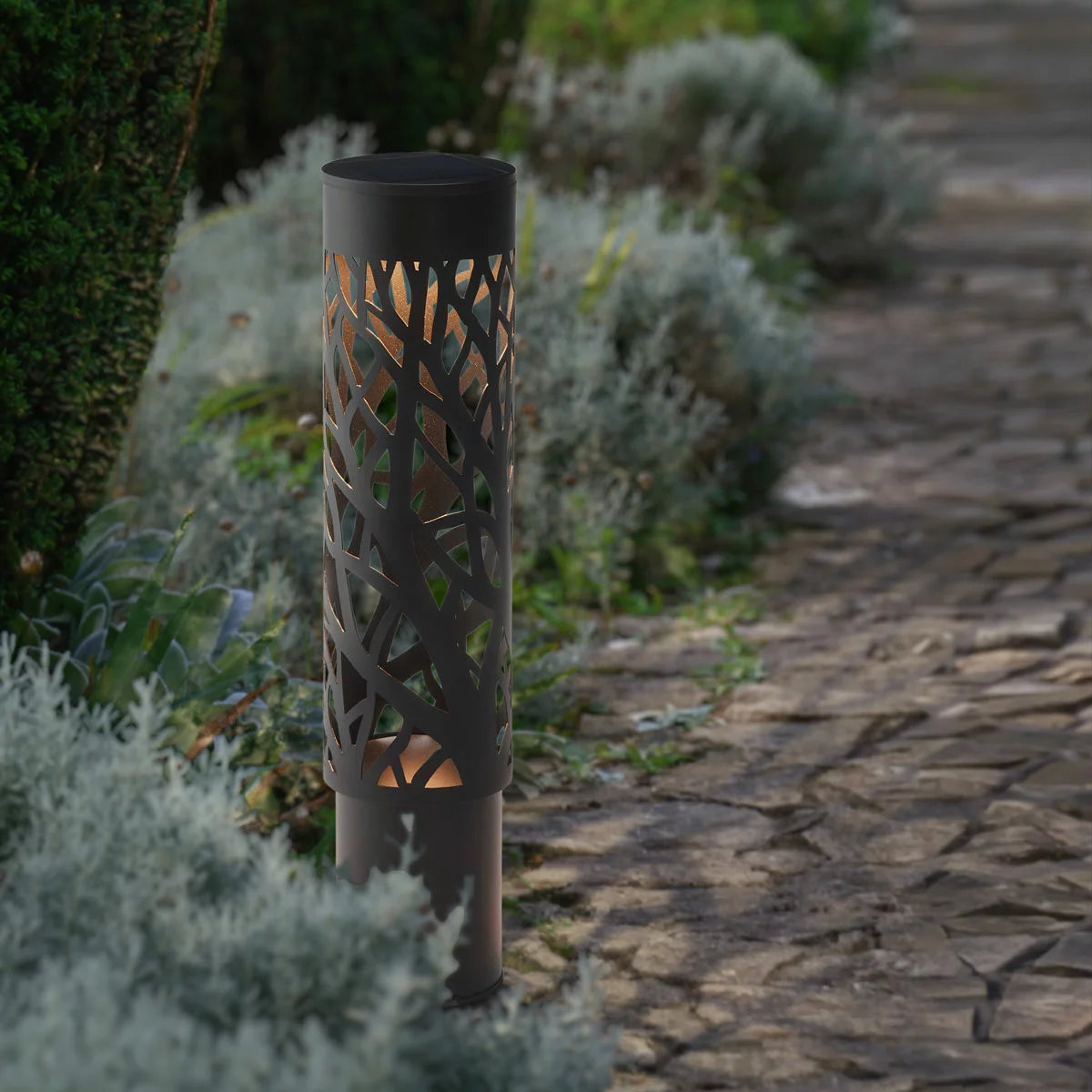 A pair of GTX steel solar bollard lights installed along a garden walkway, featuring intricate cut-out patterns that cast decorative shadows on the ground