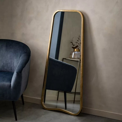 Front view of the Gallery Kurva gold metal frame leaner mirror with soft curved corners leaning against a light beige wall.