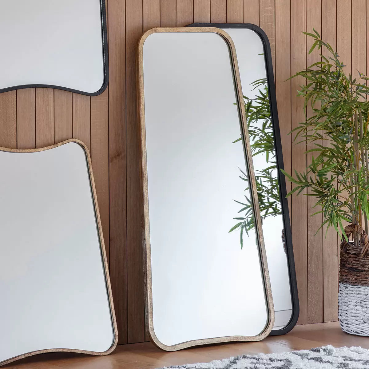 Front view of the Gallery Kurva gold metal frame leaner mirror with soft curved corners leaning against a light beige wall.