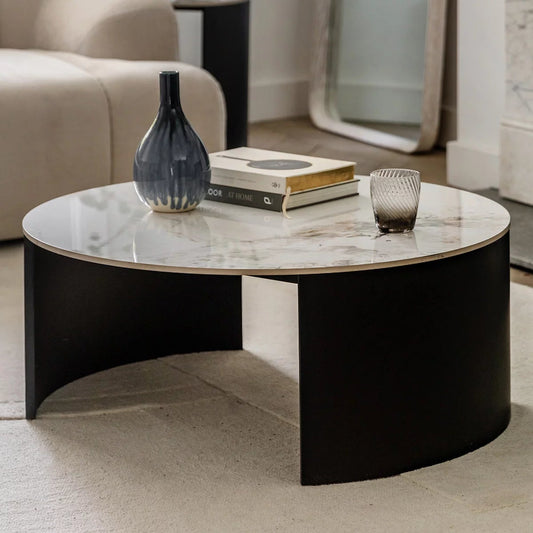 Gallery Marzio round white marble coffee table with polished Carrara stone top and curved matte black iron legs in a luxury living room.