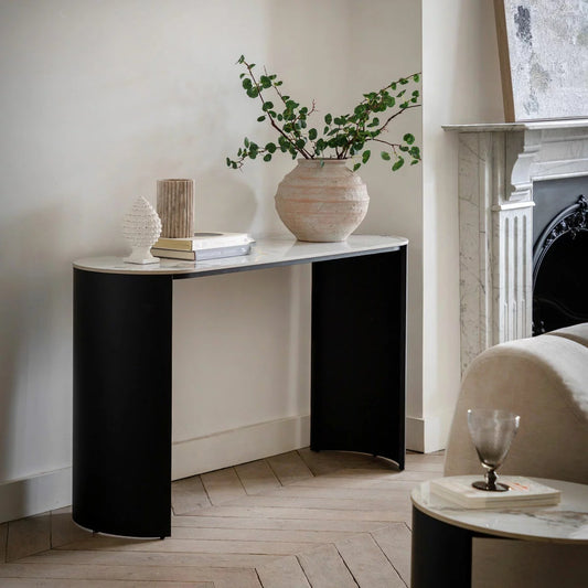 White Carrara marble style console table with a slim pill-shaped top and curved matte black metal legs, shown at a slight angle against a pale wall in a bright entryway with a vase and lamp.