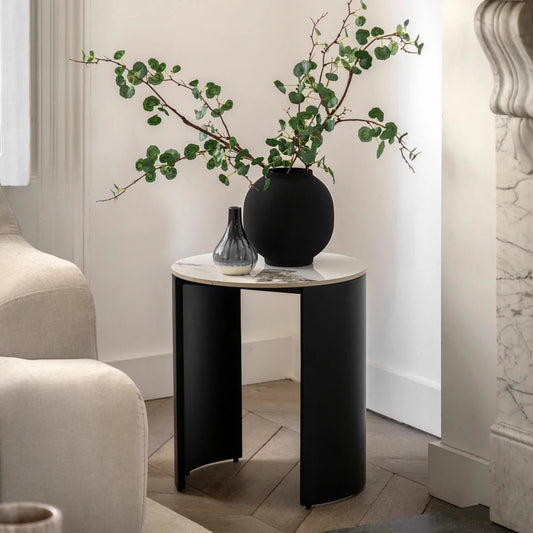 Gallery Marzio round white marble side table with Carrara stone top and curved matte black iron legs placed next to a grey fabric armchair.