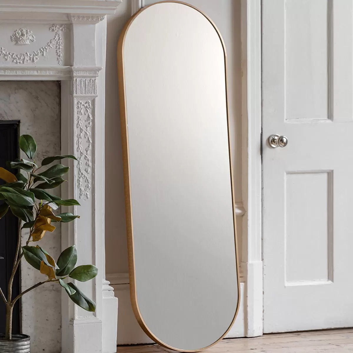 Full-length Gallery Yardley Gold Leaner Mirror with a rounded gold iron frame standing vertically in a bright modern living space reflecting natural sunlight.