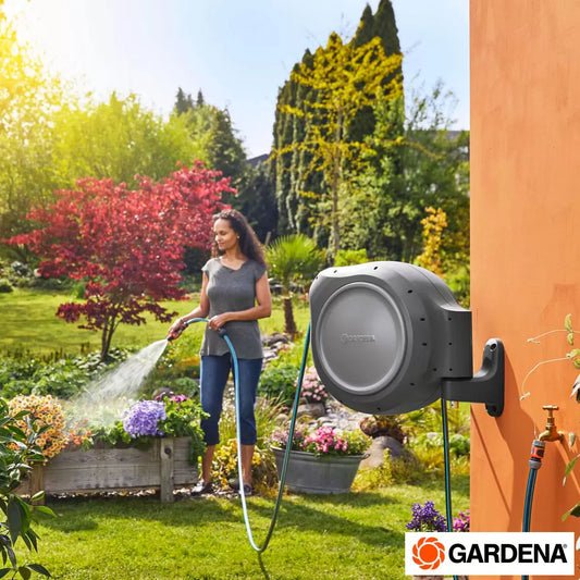 A Gardena 30m wall-mounted hose box in a clean grey and orange finish, mounted on a brick wall with the hose extended across a green garden lawn.