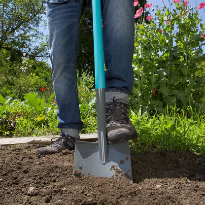 A set of Gardena ClassicLine digging spade and fork standing in a neat garden bed, accompanied by a pair of gardening gloves on a wooden potting bench.