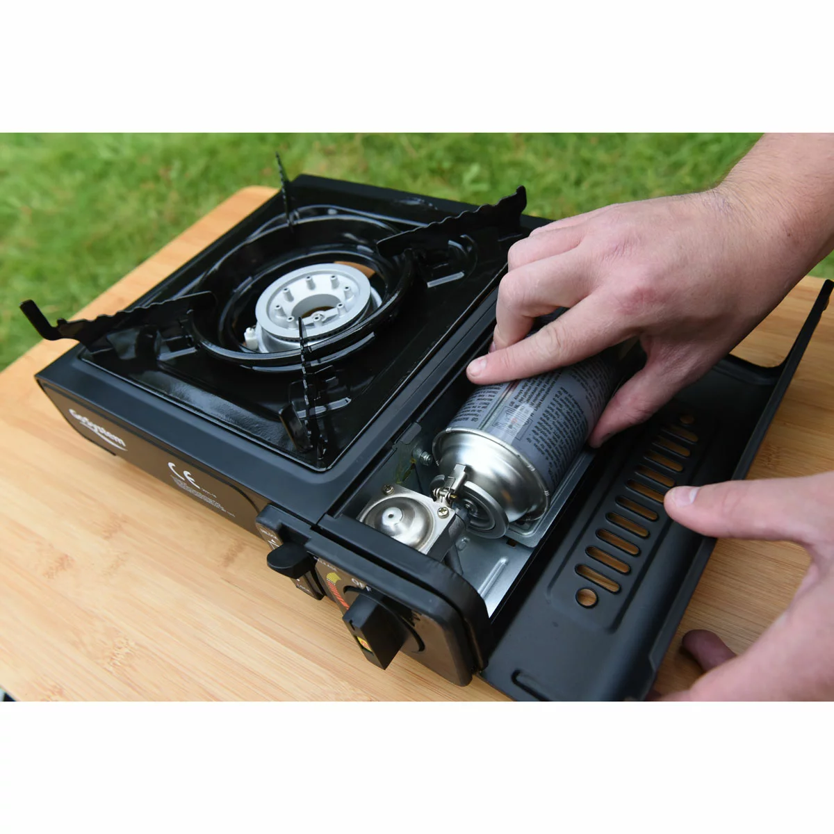 Compact black metal GoSystem Dynasty Compact II single-burner stove with serrated pan supports sitting on a wooden picnic bench next to its hardshell carrying case.