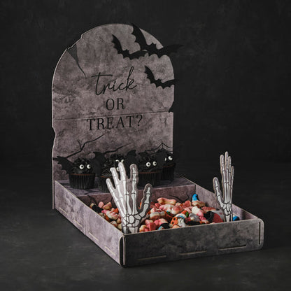 Front angle view of a grey cardboard tombstone treat stand displaying Halloween cupcakes on a dark party table.