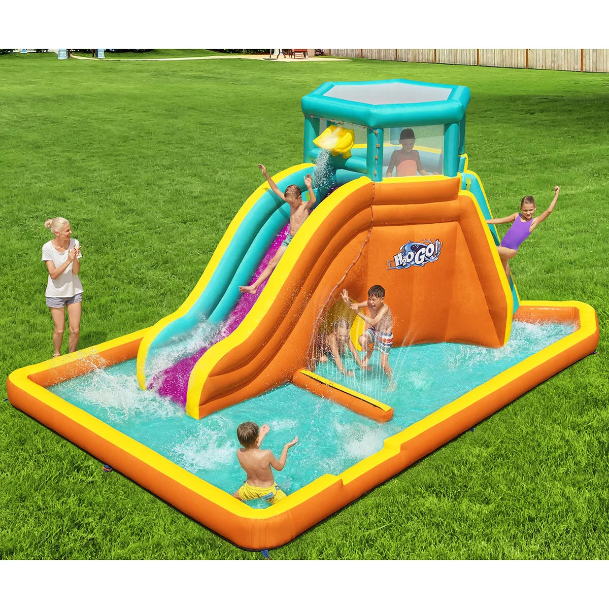 H2OGO! Tidal Tower orange inflatable mega water park with climbing tower, water slide, splash pool, crawl-through tunnel, sprinkler wall, and dump bucket set up in a sunny family garden