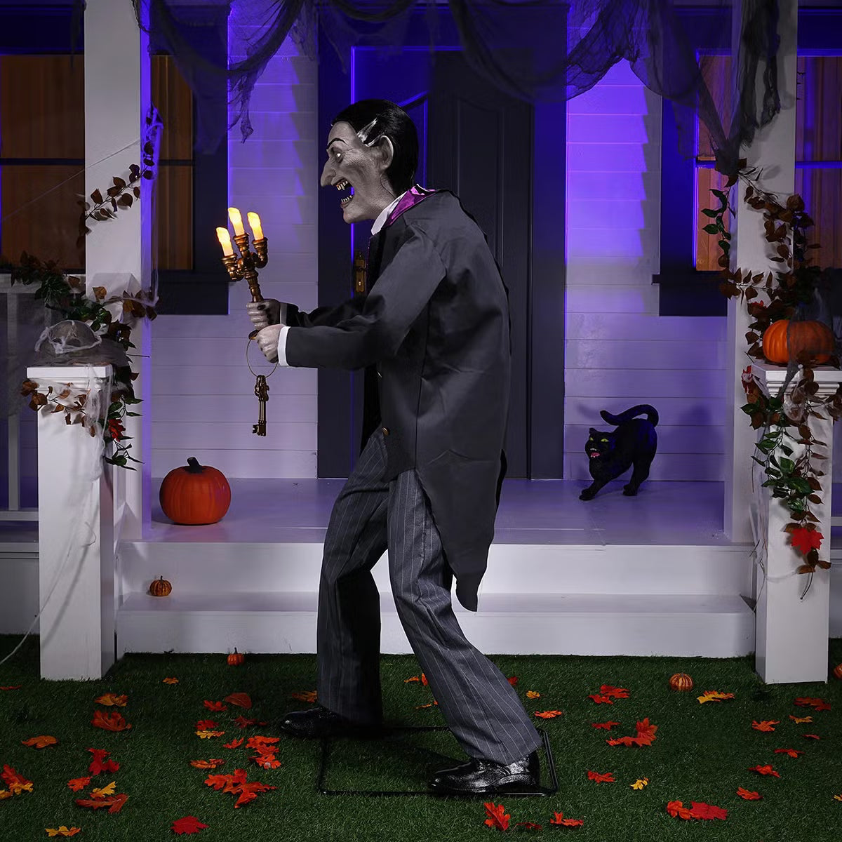 Full-length shot of a 6ft tall animated Halloween butler prop in a black suit holding an illuminated candelabra on a foggy front porch at dusk.