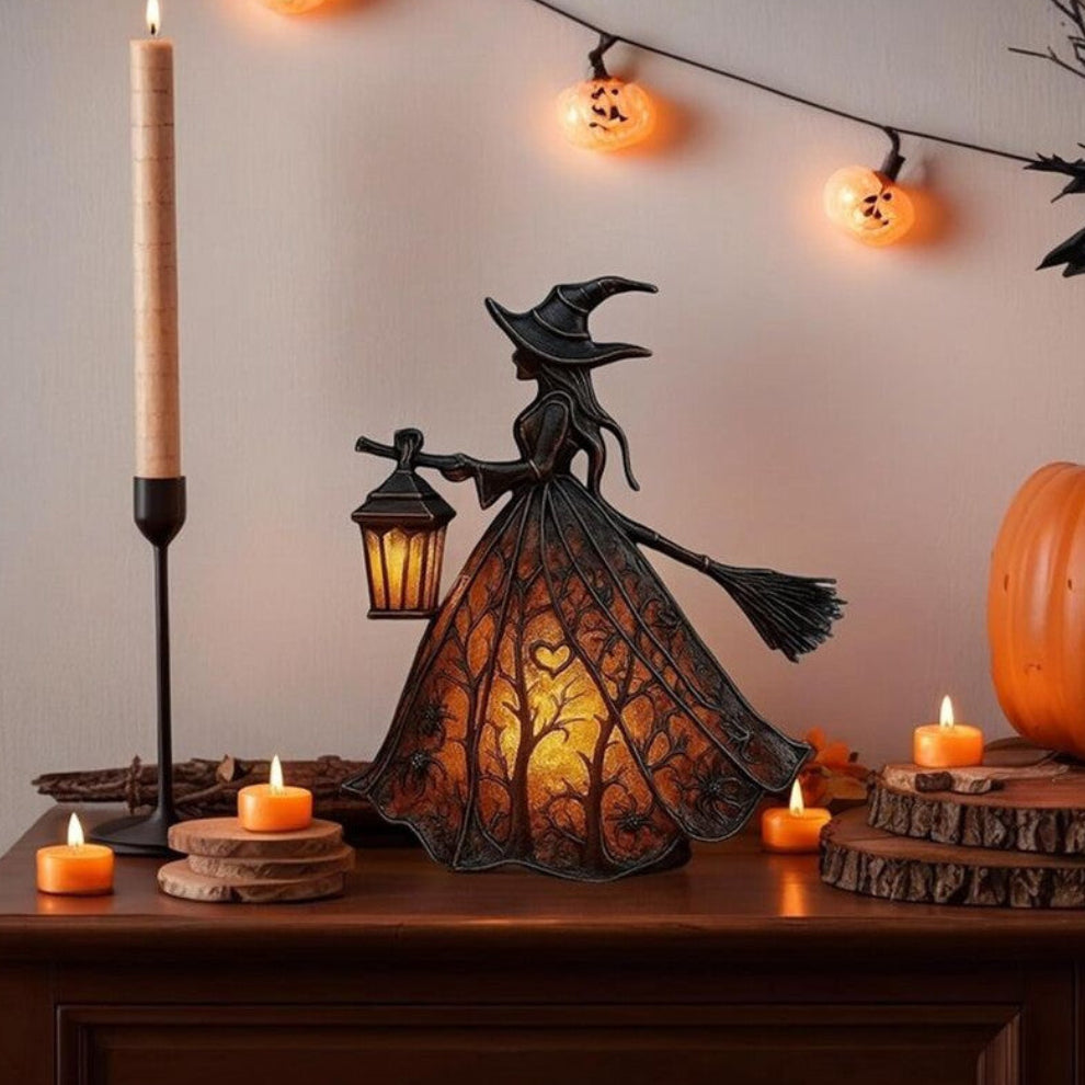 A detailed black resin Halloween gothic witch table lamp holding a glowing lantern on a dark wooden surface.