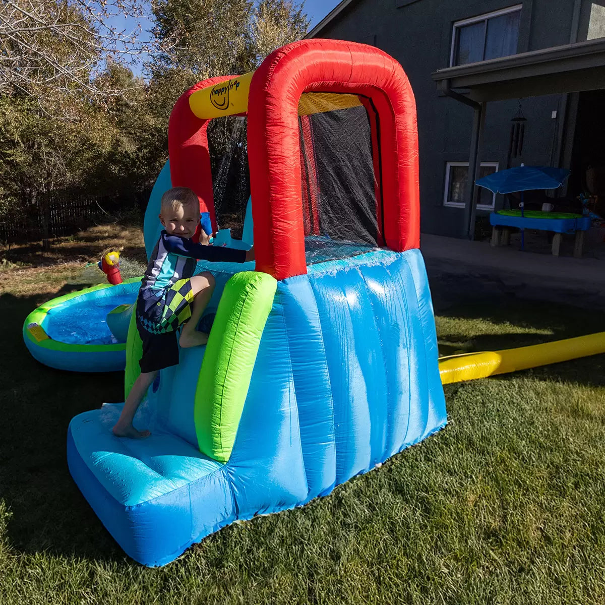 Colourful Happy Hop inflatable water slide pool with climbing wall, bounce area, splash pool and water cannon set up on a sunny garden lawn for children's outdoor summer play.