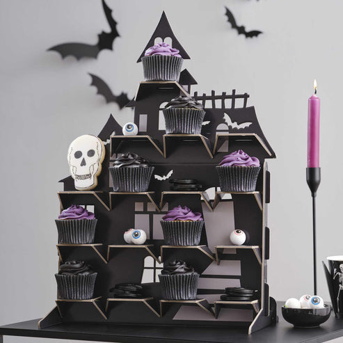 Black multi-tiered haunted house treat stand displayed on a Halloween party table filled with frosted cupcakes and dark candies.