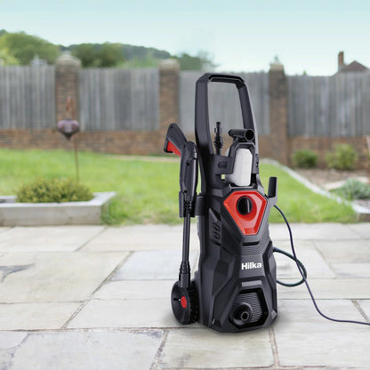 A compact Hilka 1600W pressure washer shown with its patio cleaner attachment, high-pressure nozzle, and foam bottle, set against a clean residential driveway.