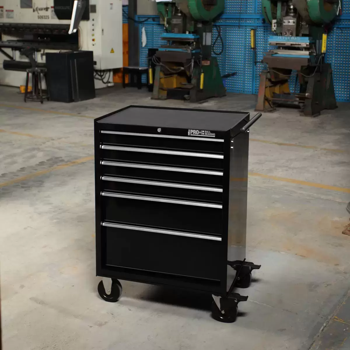 Front angle view of the black Hilka HD Pro+ 6-drawer steel tool trolley on rubber castors with drawers slightly open to show the silver aluminium pulls and black foam liners in a professional workshop environment.