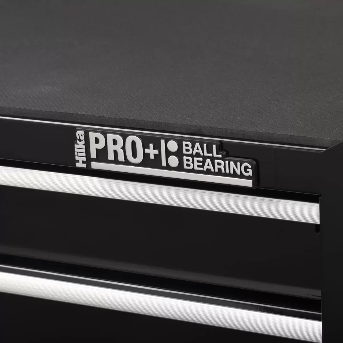 Front angle view of the black Hilka HD Pro+ 6-drawer steel tool trolley on rubber castors with drawers slightly open to show the silver aluminium pulls and black foam liners in a professional workshop environment.