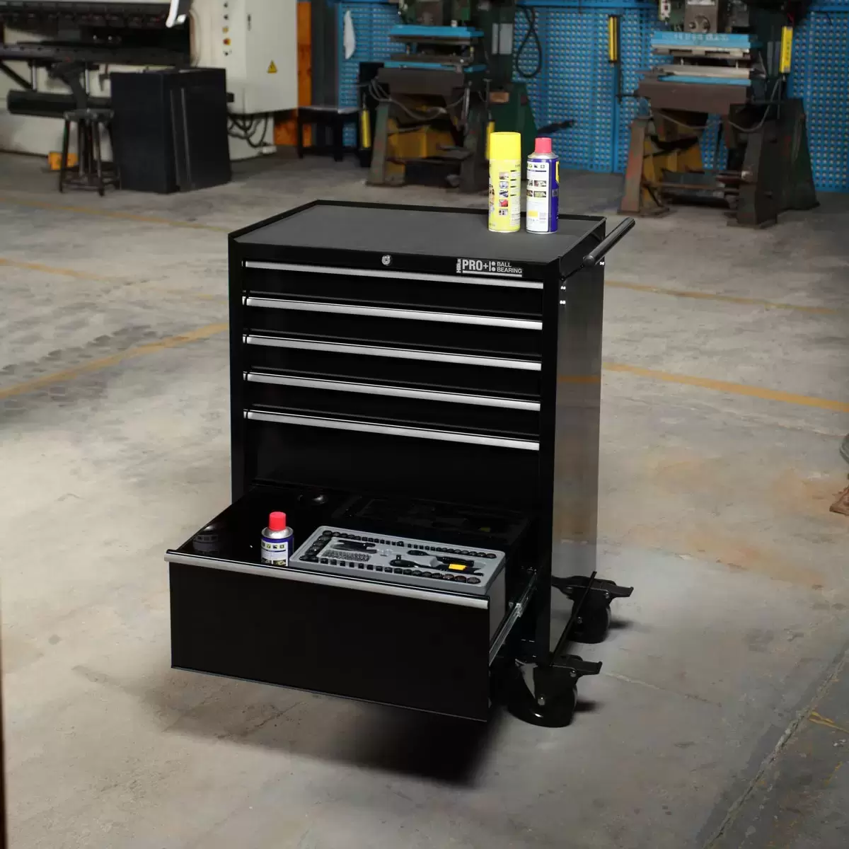 Front angle view of the black Hilka HD Pro+ 6-drawer steel tool trolley on rubber castors with drawers slightly open to show the silver aluminium pulls and black foam liners in a professional workshop environment.