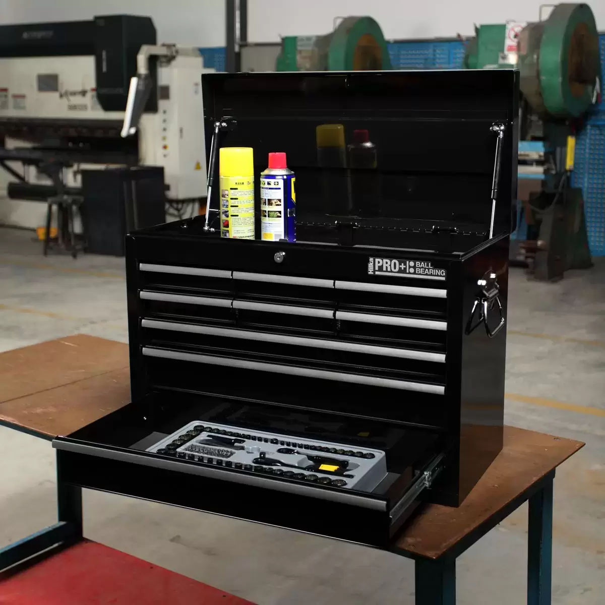 Black steel Hilka HD Pro+ 9-drawer tool chest shown from a three-quarters angle with the top lid open on dual gas struts, showcasing the aluminium drawer pulls and red interior details on a rustic wooden workbench.