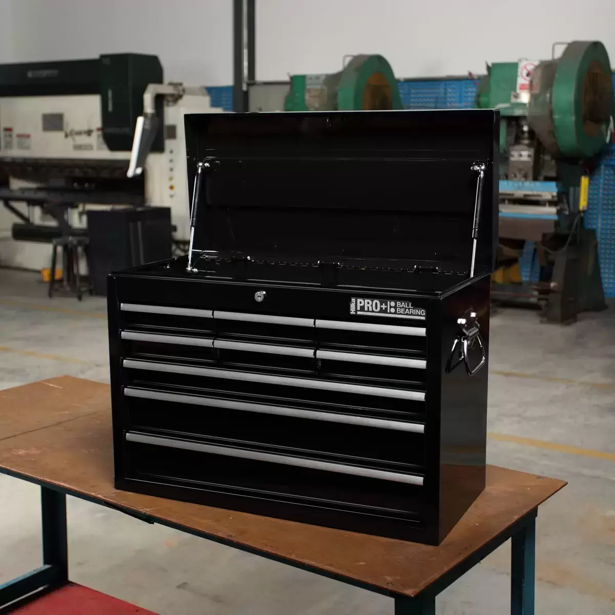 Black steel Hilka HD Pro+ 9-drawer tool chest shown from a three-quarters angle with the top lid open on dual gas struts, showcasing the aluminium drawer pulls and red interior details on a rustic wooden workbench.