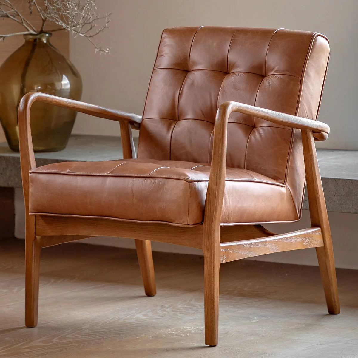 Side profile of the Gallery Humber Armchair featuring rich brown tufted top-grain leather cushions set against a warm, angled solid oak wooden frame in a brightly lit modern living room.