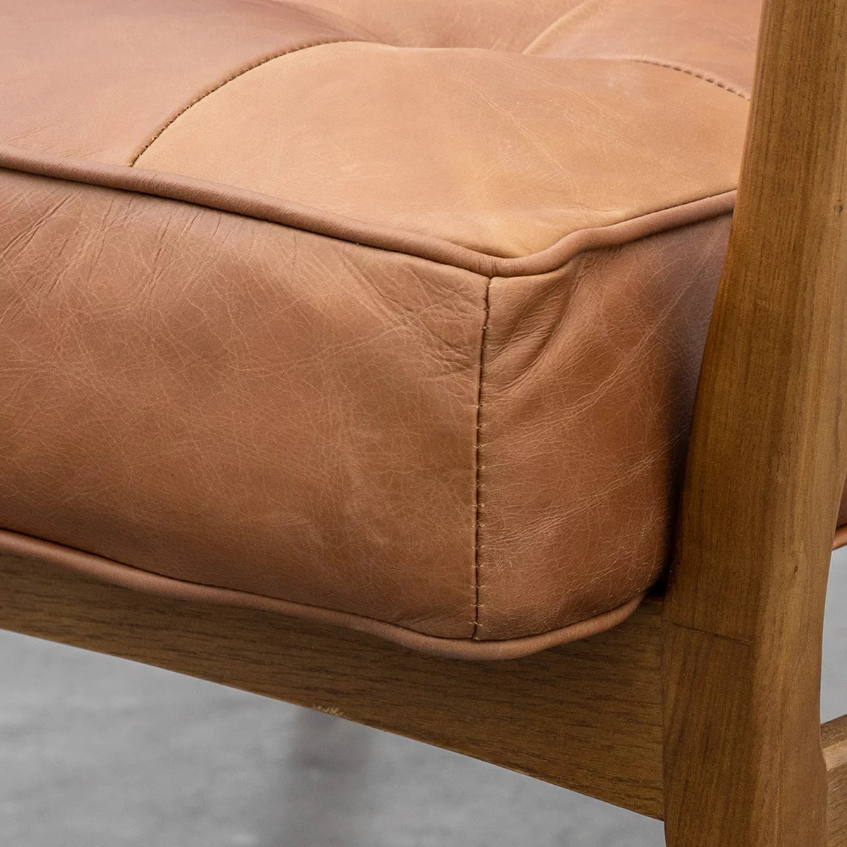 Side profile of the Gallery Humber Armchair featuring rich brown tufted top-grain leather cushions set against a warm, angled solid oak wooden frame in a brightly lit modern living room.