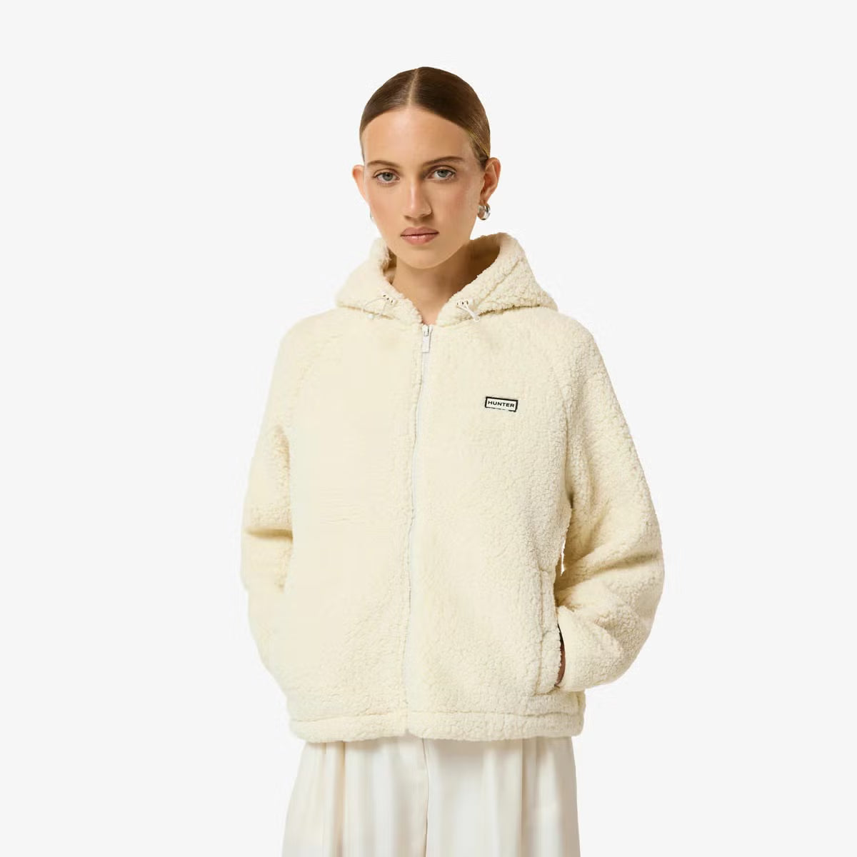 Side angle view of a woman wearing a beige Hunter Women's Highland Hooded Fleece with plush borg texture and a zip-up hood outdoors.